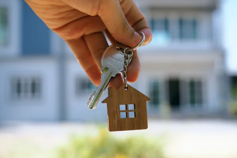 A hand holding a house key and a keychain shaped like a small house, with a blurred building in the background.